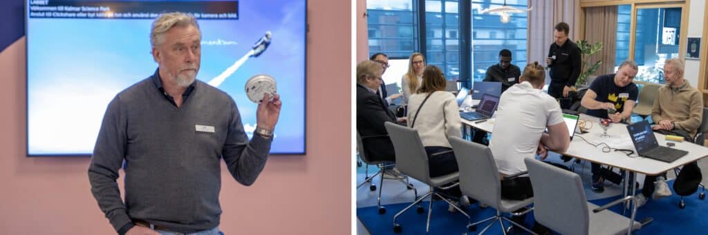 En person håller upp en teknisk enhet framför en stor presentationsskärm. Bredvid syns en grupp människor som sitter runt ett konferensbord och arbetar tillsammans med datorer och teknisk utrustning.