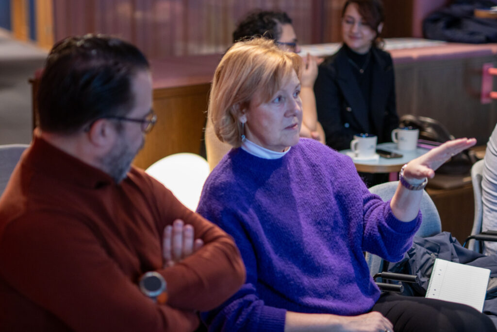 Deltagare sitter i mindre grupp under ett event. En person i lila tröja gestikulerar medan andra lyssnar. Kaffekoppar och anteckningar syns på borden.